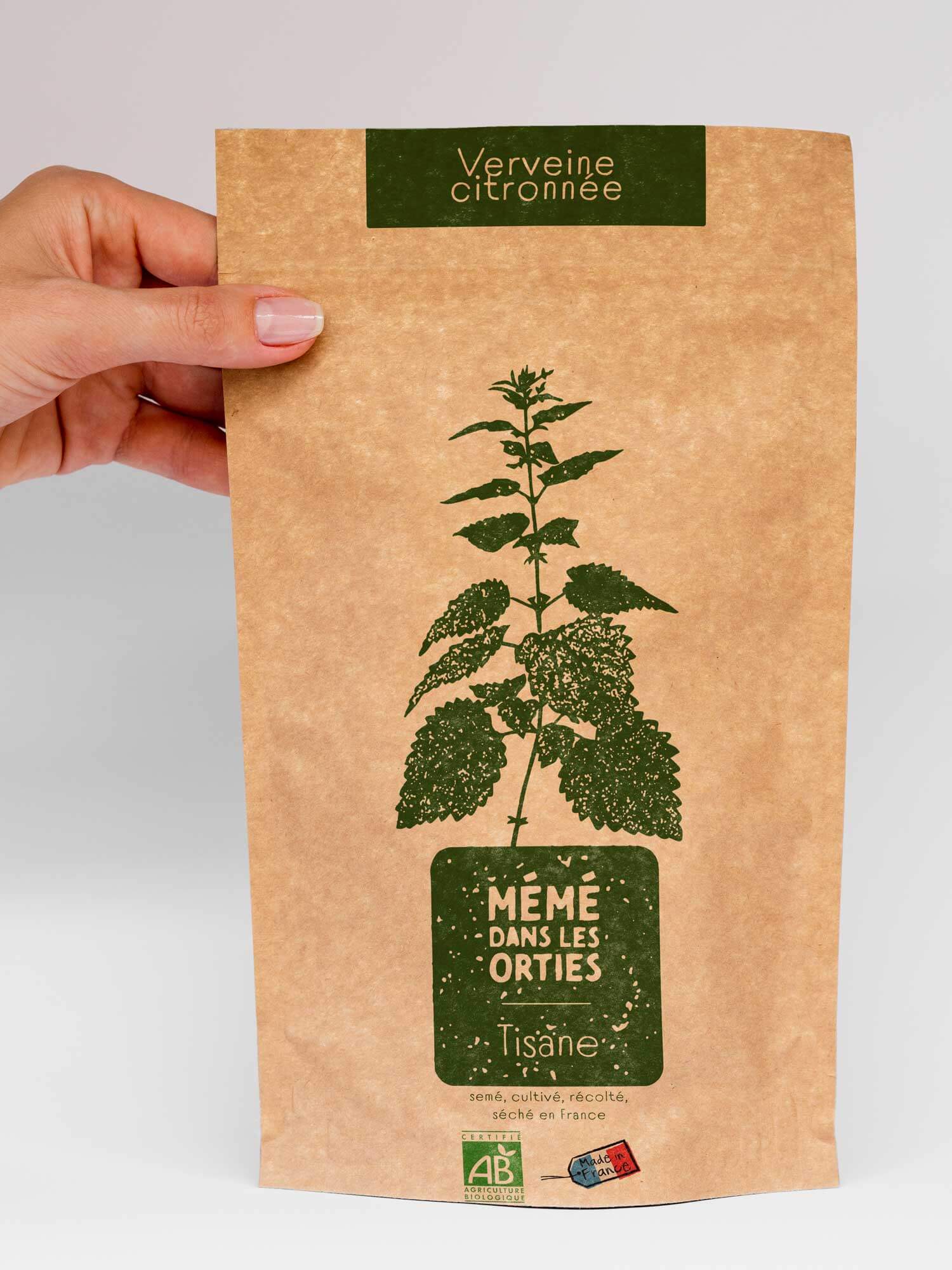 Photographie d’un sachet kraft de tisane tenu par une main. Packaging réalisé par le studio nueva vista. Sur le haut du packaging, un encadré vert foncé indique « Verveine citronnée ». Au centre, une illustration végétale verte représente la plante. En dessous, un cartouche vert porte le nom de la marque « Mémé dans les orties » suivi du mot « Tisane ». En bas du sachet figurent les mentions « semé, cultivé, récolté, séché en France » ainsi que deux labels : « AB – Agriculture Biologique » et un petit macaron « Made in France ».