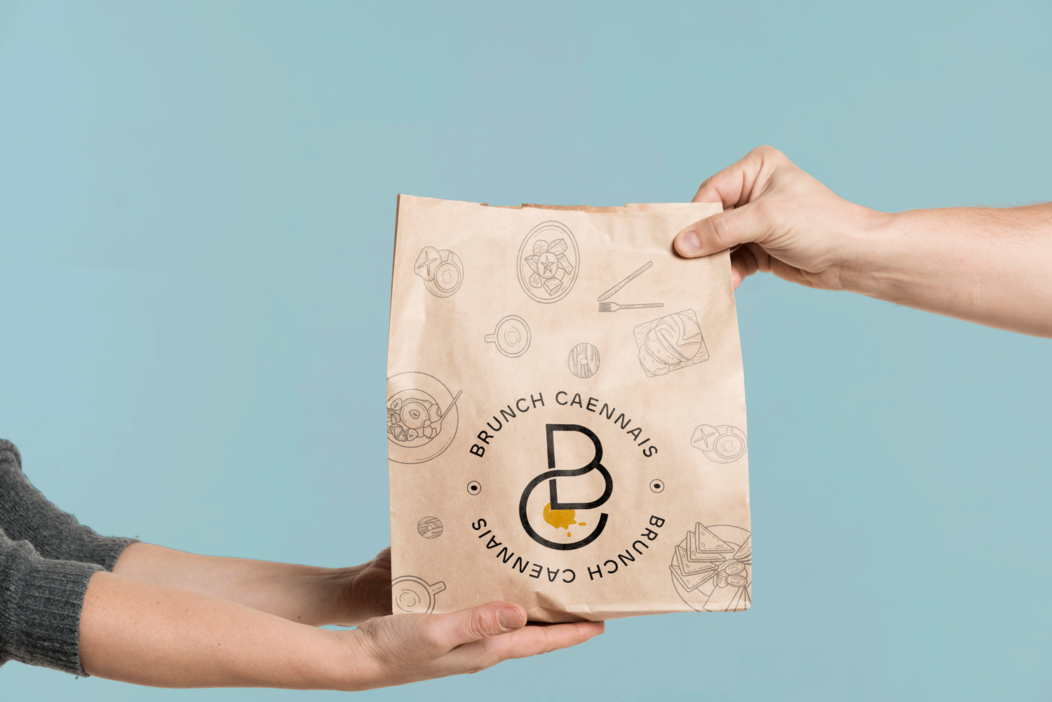 Sachet kraft tenu à deux mains, présentant le logo “Brunch Caennais” conçu par Gaële Gerbaux du studio nueva vista : monogramme circulaire “BC” avec touche jaune et illustrations fines de plats de brunch imprimées sur le sac, sur fond bleu clair.