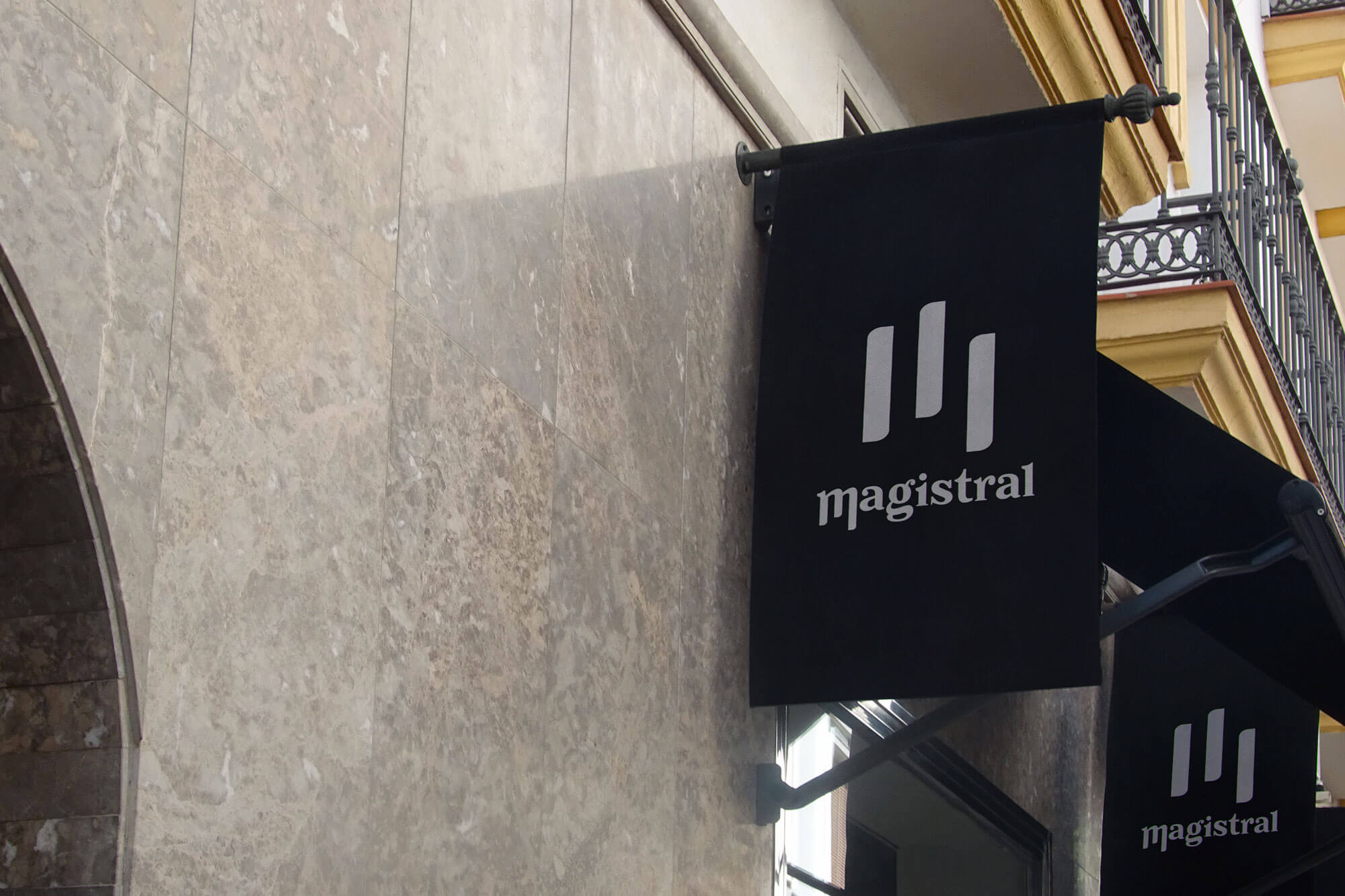Enseigne drapeau noire affichant le logo “magistral” conçu par Gaële Gerbaux du studio nueva vista pour un magasin d’instruments de musique : symbole vertical évoquant des touches de piano et lettrage typographique blanc, installés sur une façade en pierre.