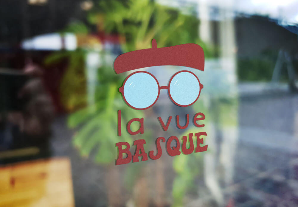 Enseigne adhésive sur vitrine présentant le logo “La Vue Basque” pour un opticien, conçu par Gaële Gerbaux du studio nueva vista : lunettes rondes bleu clair surmontées d’un béret basque stylisé, accompagnées d’un lettrage rouge évoquant l’identité locale.