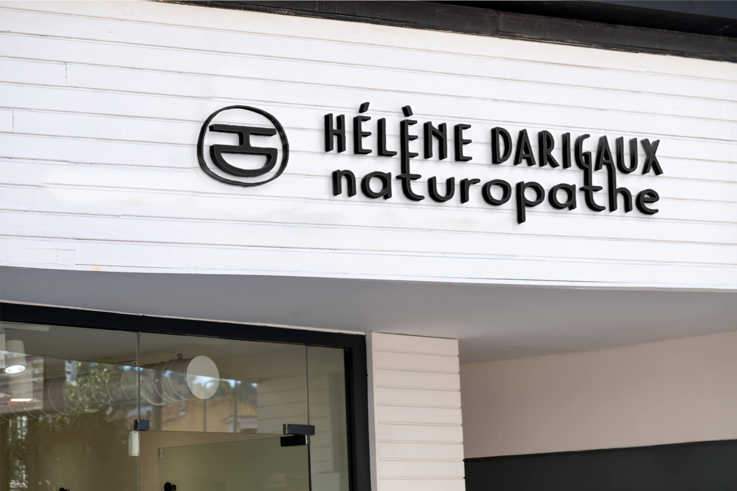 Enseigne extérieure d’un cabinet de naturopathie présentant le logo et le lettrage “Hélène Darigaux naturopathe” conçus par Gaële Gerbaux du studio nueva vista : symbole arrondi en 3D et typographie noire installés sur un bardage bois blanc.