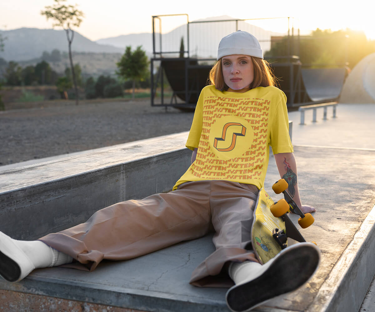 Mockup photo d’une rideuse assise dans un skatepark au coucher du soleil, portant un t-shirt jaune avec le logo “System” conçu par Gaële Gerbaux du Studio nueva vista (concepteur d’espaces de sports urbains — skatepark, pumptrack, streetpark). Le design du t-shirt met en avant le logo dans un cercle central et un motif répétitif en arrière-plan, présenté dans un contexte sportif réaliste.